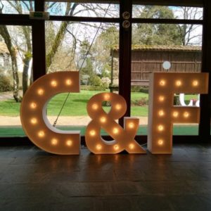 Letras gigantes 'C&F' de Animus Discotecas iluminando con bombillas un evento, con vistas al jardín verde desde el interior.