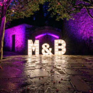 Mágicas letras gigantes 'M&B' iluminando la entrada de un evento nocturno, perfectas para un matrimonio inolvidable.